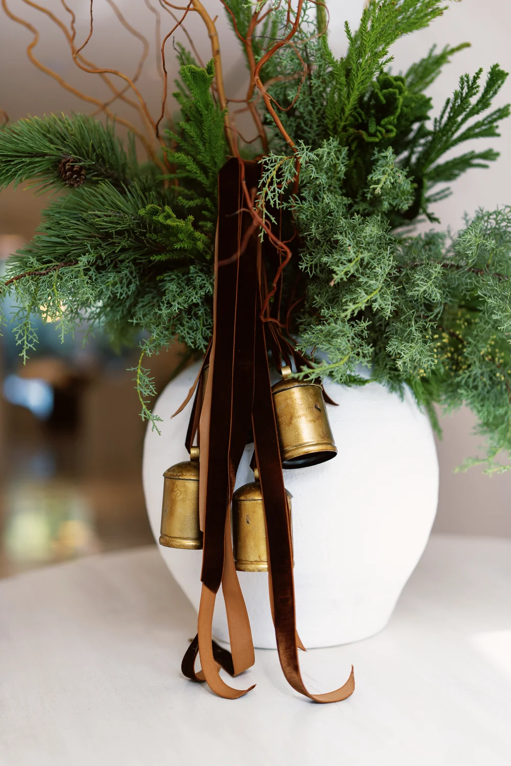 Evergreen Christmas centrepiece in a white vase with brown velvet ribbons and brass bells