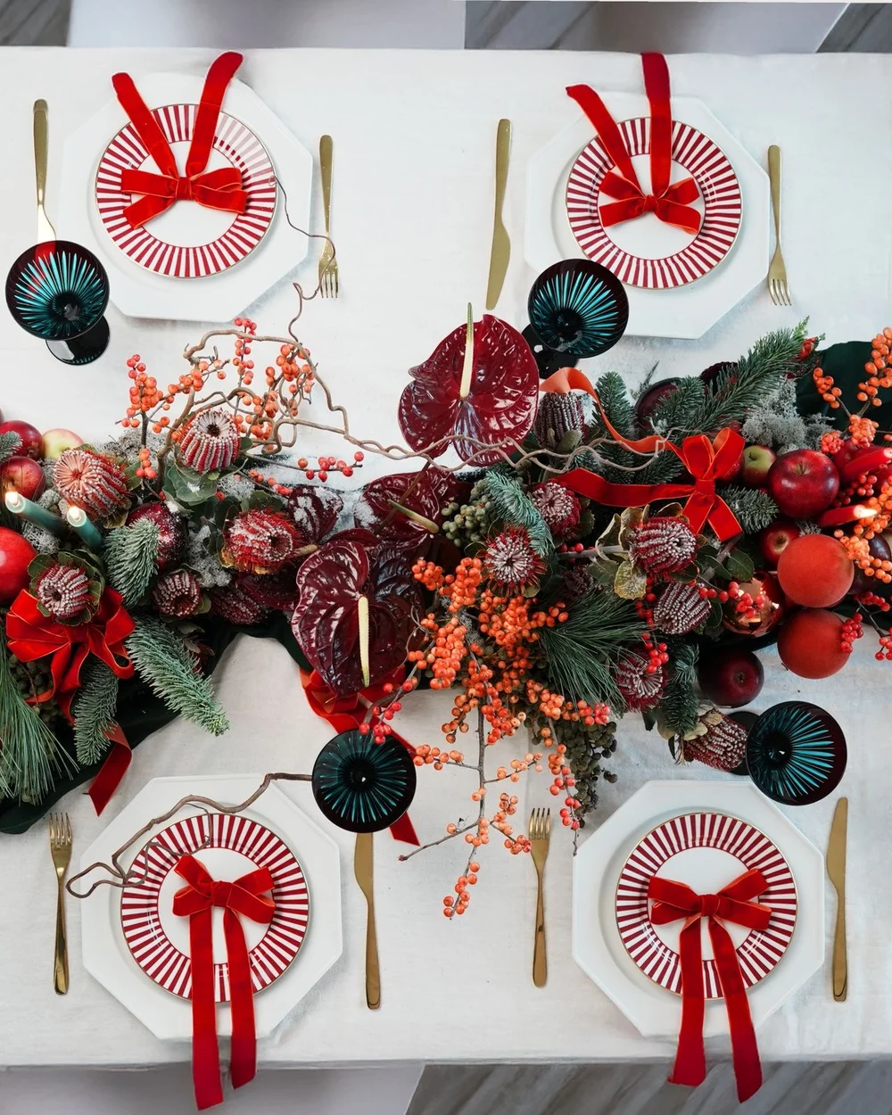 Festive dining table with red ribbon place settings, dark glassware and an evergreen, berry and burgundy anthurium centrepiece