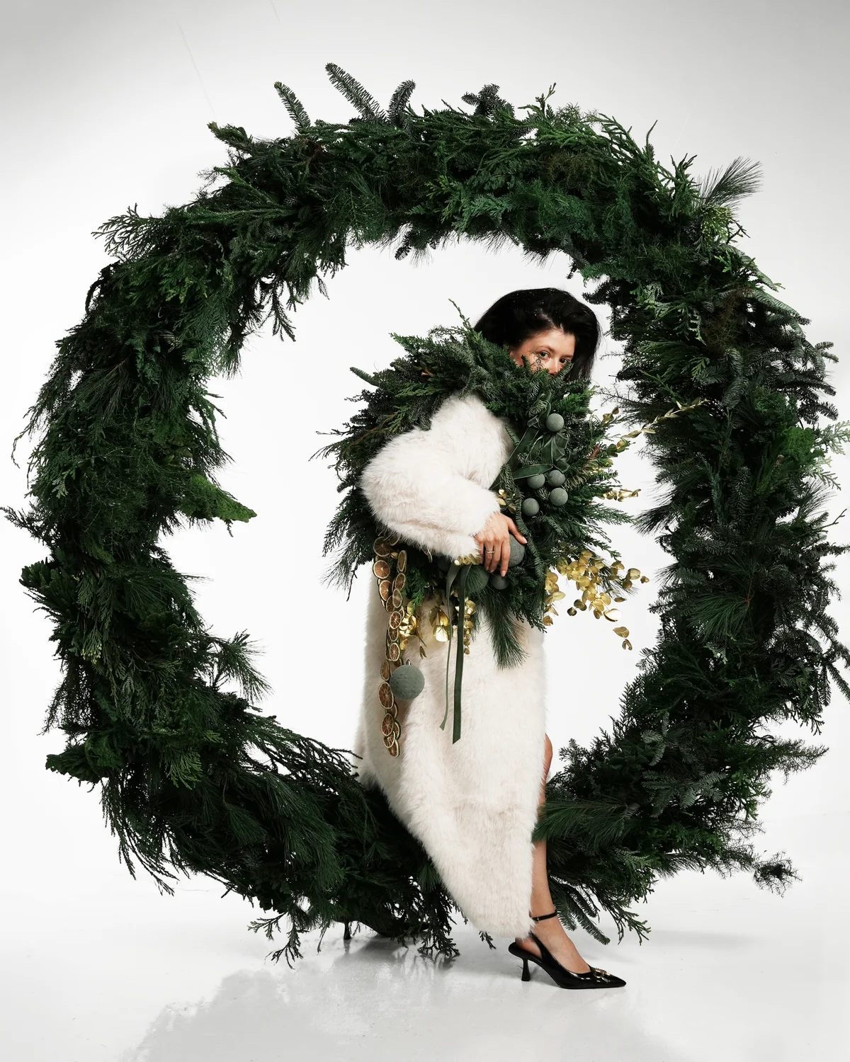 Woman framed by a large circular evergreen wreath, wearing festive foliage and gold accents