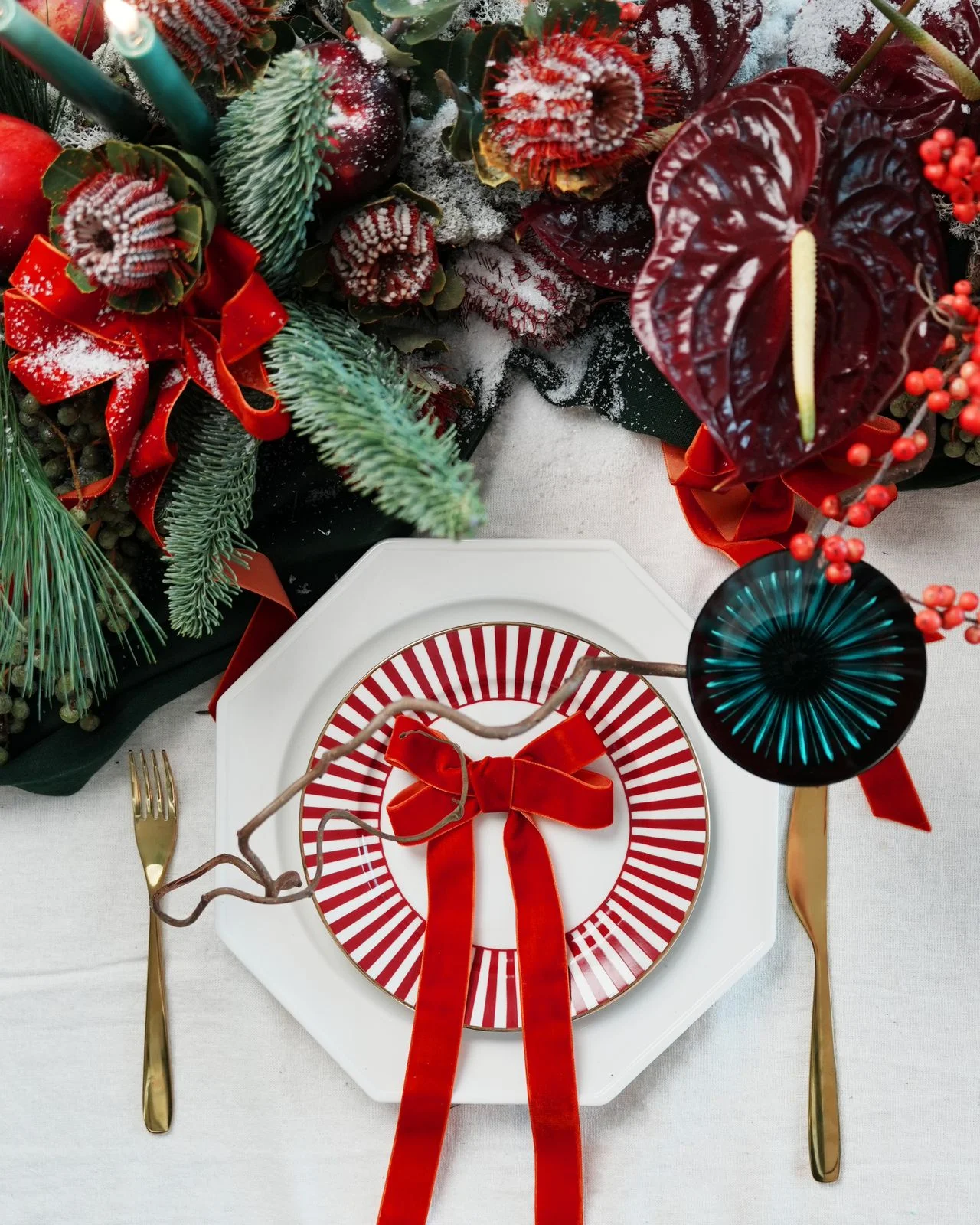 Christmas table setting with a red-striped plate, red velvet bow, gold cutlery, evergreen foliage and burgundy flowers
