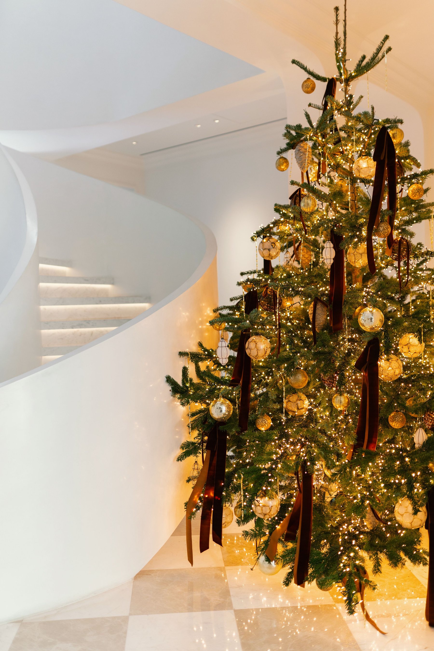 Christmas tree with golden ornaments, burgundy ribbons and warm lights beside a curved white staircase
