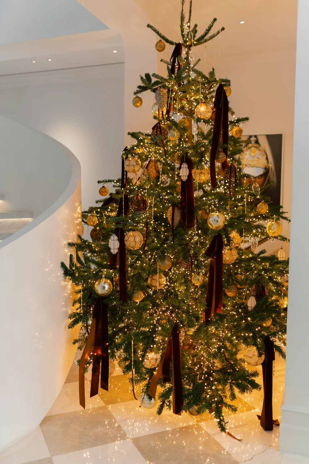 Christmas tree with warm white lights, gold ornaments and burgundy velvet ribbons beside a curved white staircase