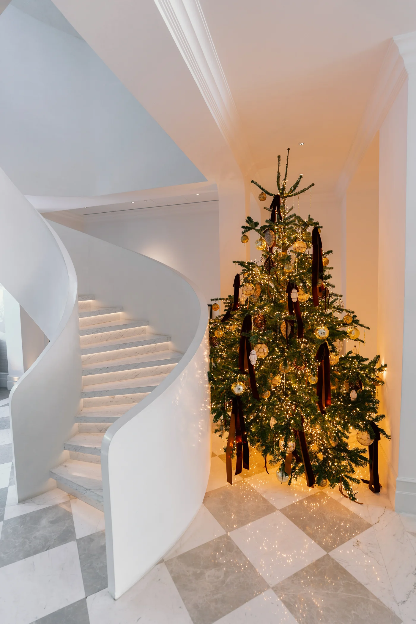 Christmas tree with warm lights, gold ornaments and burgundy ribbons beside a curved white staircase